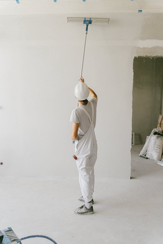 Back view of a painter in overalls and hard hat painting an interior wall with a roller.
