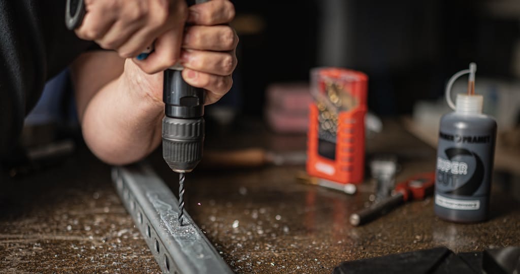 Hands using a power drill on metal, surrounded by workshop tools and equipment.